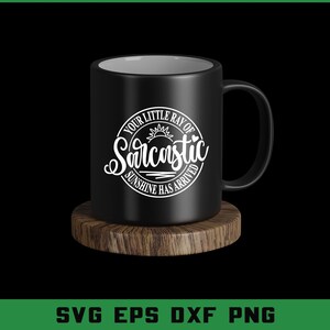 Puede incluir: Una taza de cer&aacute;mica negra con interior blanco y asa, sobre un posavasos de madera. La taza presenta un dise&ntilde;o circular con el texto "Your Little Ray of Sarcastic Sunshine Has Arrived."