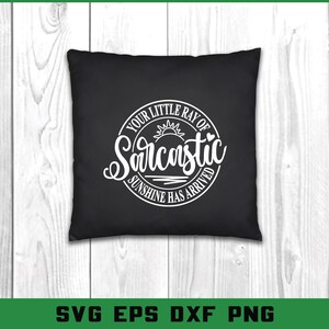 Puede incluir: Coj&iacute;n cuadrado negro con texto blanco que dice "YOUR LITTLE RAY OF Sarcastic SUNSHINE HAS ARRIVED" en un dise&ntilde;o circular. El dise&ntilde;o incluye un sol y un coraz&oacute;n. El coj&iacute;n est&aacute; sobre una superficie de madera clara.
