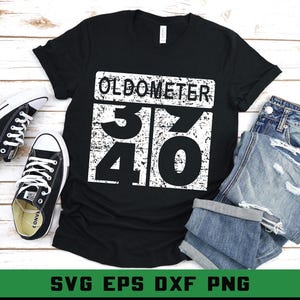 May include: Black t-shirt with the word "OLDOMETER" and the numbers "37" and "40" in a white, distressed font. The shirt is paired with blue jeans and black canvas trainers. The image is on a wooden surface.