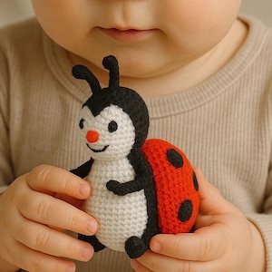 Pode incluir: Um bebê segura um brinquedo de joaninha de crochê. O brinquedo tem uma concha vermelha com manchas pretas, um corpo branco, antenas e braços pretos e um rosto sorridente. O bebê está vestindo uma camisa de manga comprida marrom claro.