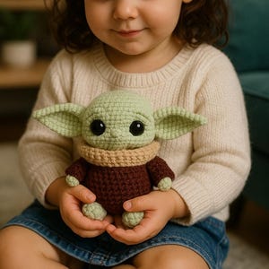 Puede incluir: Muñeco de ganchillo con la imagen de un personaje popular, con cabeza y orejas verde claro, ojos negros grandes y cuerpo marrón. El muñeco lo sostiene un niño que lleva un jersey color crema y una falda vaquera.
