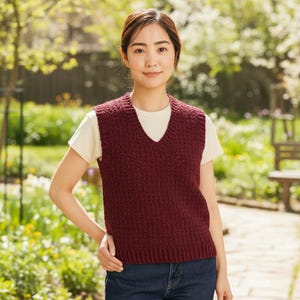 May include: A burgundy knitted vest with a V-neck, worn over a white t-shirt. The vest has a textured pattern and a ribbed hem. The person is standing outside in a garden setting, wearing blue jeans.