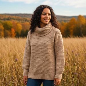 May include: A tan, textured knit sweater with a cowl neck. The sweater has ribbed cuffs and a ribbed hem. The person in the image is wearing blue jeans and is standing in a field of tall grass with a background of trees.