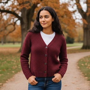 Op de afbeelding: Een bordeauxrode cardigan met een textuurgebreid patroon en een V-hals. De cardigan heeft drie donkere knopen aan de voorkant en wordt gedragen over een wit t-shirt en een blauwe spijkerbroek. De achtergrond toont een park met bomen en een wandelpad.
