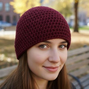 May include: A burgundy crochet beanie hat. The hat is made of a textured, knitted material and is worn on the head. The background is blurred, showing a park setting with trees and a bench.