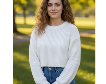 Könnte beinhalten: Eine Frau trägt einen weißen, gecroppten Pullover und blaue Jeans. Der Pullover hat eine gerippte Strickstruktur und lange Ärmel. Die Frau hat lockiges braunes Haar und steht draußen vor einem verschwommenen grünen Hintergrund.