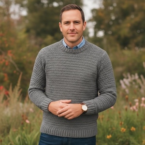 Könnte beinhalten: Ein grauer Pullover mit Rundhalsausschnitt und strukturiertem Strickmuster. Der Pullover hat lange Ärmel und gerippte Bündchen und Saum. Die Person trägt eine Uhr mit braunem Lederarmband. Der Hintergrund ist eine verschwommene Szene im Freien.