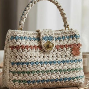 Pode incluir: Bolsa de mão de crochê feita à mão, na cor bege com listras horizontais azuis, verdes e ferrugem. Possui uma alça trançada, um fecho em forma de flor e uma pequena etiqueta de couro. A bolsa tem aproximadamente 25 cm de largura e 20 cm de altura.