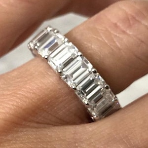 May include: A close-up of a silver ring with multiple rectangular-cut, clear gemstones set in a row. The ring is on a finger, with the skin tone visible in the background.