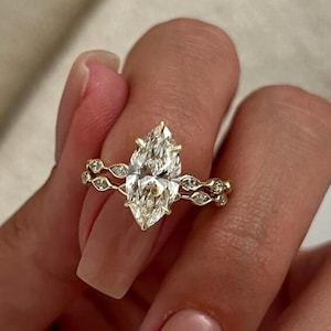 May include: A gold engagement ring featuring a marquise-cut diamond. The band has a delicate design with small accent stones. The ring is displayed on a hand with manicured nails.