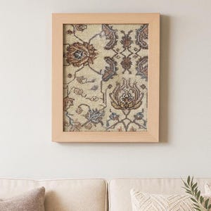 May include: A framed wall art piece featuring a section of a patterned rug. The rug design includes floral and geometric elements in shades of brown, blue, and cream. The frame is made of light-colored wood.