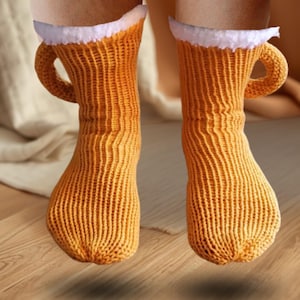 Könnte beinhalten: Ein Paar neuartige Socken, die Bierkrügen nachempfunden sind. Die Socken sind in einem warmen Orangeton gehalten und haben einen weißen, flauschigen Rand am oberen Rand sowie einen gebogenen Griff an der Seite. Die Socken sind gestrickt.