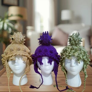 May include: Three handmade crochet hats in tan, purple, and green, each with a unique spiky design on top. The hats have ear flaps and ties, displayed on white mannequin heads. The hats are a whimsical and creative accessory.