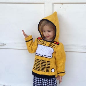 Puede incluir: Un suéter amarillo de ganchillo con capucha con un diseño de autobús escolar. El suéter tiene ribetes negros y las palabras "School Bus" están bordadas en la parte delantera.