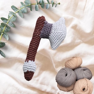May include: A crocheted axe toy in shades of brown, gray, and white, with a textured appearance. The axe head features a gradient from gray to white. Several balls of yarn in neutral tones are also visible.