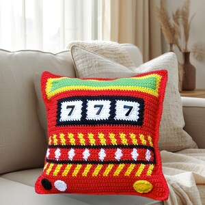 May include: A square, red crochet pillow with a slot machine design. The pillow features the number "7" three times in white, a green and yellow top border, and yellow and white diamond patterns. Black and yellow accents complete the design.