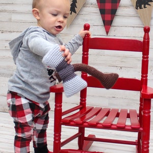 May include: A toddler wearing red and white plaid pants and a gray hooded sweatshirt holds a crocheted toy axe. The toddler is standing in front of a red rocking chair with a white wood background.
