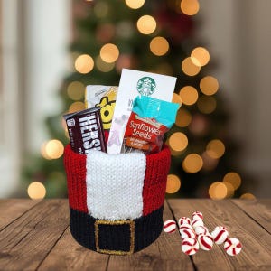 May include: A festive gift basket shaped like Santa's belt, filled with snacks and a Starbucks gift card. The basket is red, white, and black with a gold buckle. Peppermint candies are scattered on the wooden surface.