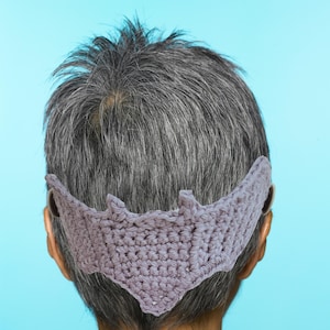 May include: A crocheted bat-shaped headband in a light purple colour. The headband is worn on the back of the head, covering the ears.