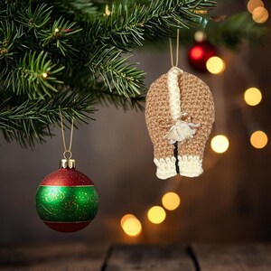 May include: Two Christmas ornaments hanging from a tree. One is a red and green bauble with gold detailing. The other is a brown and cream crocheted ornament with a bow and white accents. Festive holiday decor.