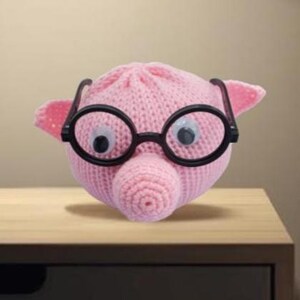 May include: A pink knitted pig with large googly eyes and black-rimmed glasses. The pig has small ears and a spiral-shaped snout. The toy is sitting on a wooden surface.
