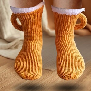 May include: A pair of novelty socks designed to resemble beer mugs. The socks are a warm orange color with a white, fluffy trim at the top, and a curved handle on the side. The socks are made of a knitted material.
