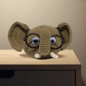 May include: A crocheted elephant head with large blue eyes, white tusks, and black-rimmed glasses. The elephant is a light brown color and has large, floppy ears. It is sitting on a wooden surface.