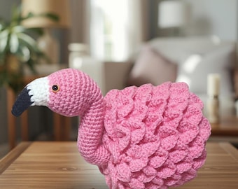 May include: A crocheted flamingo figure featuring a bright pink body with layered, textured yarn feathers, a long curved neck, and a white head with a black-tipped beak. The flamingo has a single visible dark eye with a yellow iris.