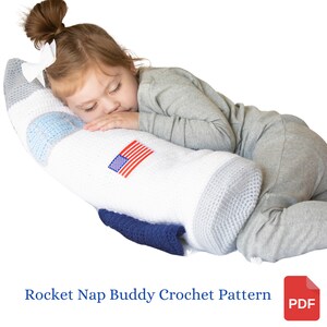 Puede incluir: Un compañero de siesta en forma de cohete tejido en blanco y gris con un diseño de estrella azul y blanca y una bandera estadounidense en el lateral. Un niño pequeño está durmiendo con la cabeza apoyada en el cohete.