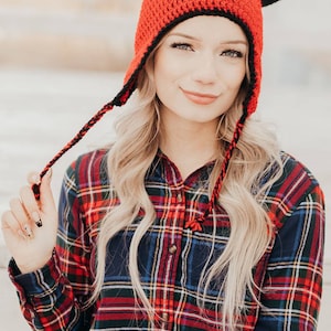 May include: A woman wearing a red and black crocheted hat with horns. The hat has a long, black tassel. She is wearing a plaid shirt.