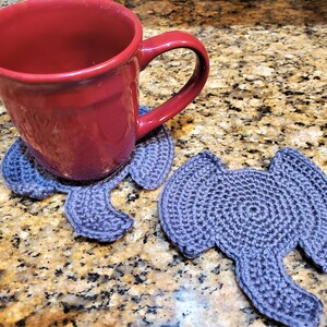 May include: Two gray crocheted elephant-shaped coasters with a red ceramic mug sitting on one of them. The coasters are on a granite countertop.