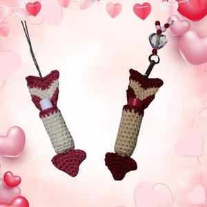 Könnte beinhalten: Zwei handgefertigte Lippenbalsamhalter aus Häkelarbeit im Valentinstag-Thema. Die Halter sind pfeilförmig, mit bordeauxfarbenem und cremefarbenem Garn. Einer hat eine Kette, der andere einen Perlenanhänger. Der Hintergrund ist rosa mit Herzdekorationen.