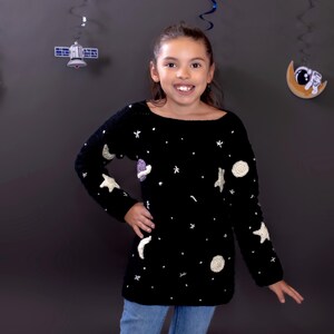 May include: A black sweater with a crew neck, adorned with white stars, moons, and other space-themed appliqués. The sweater is worn with blue jeans. Decorative space-themed hanging ornaments are in the background.