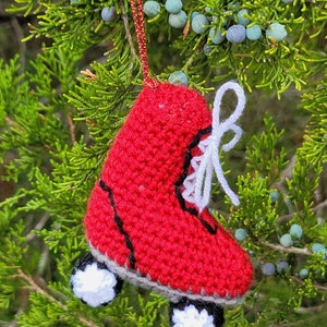 May include: Red crocheted roller skate ornament with white laces and white wheels. The ornament is hanging from a red string on a green tree branch.