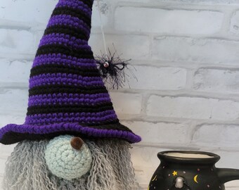 May include: A crocheted gnome wearing a tall purple and black striped pointed hat with a small dangling spider, featuring a light blue round nose and long gray yarn hair, positioned next to a black ceramic mug painted with yellow moon and fire motifs.