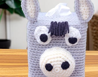 May include: A light gray crocheted tissue box cover shaped like a donkey. It features two upright ears with white centers, a white muzzle with two black circular nostrils, two white eyes with black pupils, and a dark gray yarn fringe for hair.
