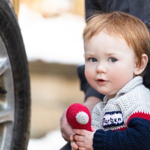 Może przedstawiać: Małe dziecko z czerwonymi włosami trzyma czerwoną i białą pluszową zabawkę. Dziecko nosi szary, czerwony i niebieski sweter na drutach z imieniem "Merten" wyszytym na rękawie.