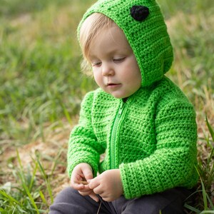 May include: A bright green, crocheted baby hoodie with a frog design, featuring a hood with small ears and black eye accents. The jacket has a zipper closure. The child is wearing gray pants and is sitting in the grass.