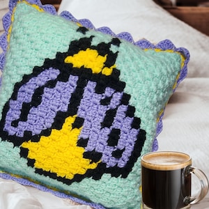 May include: A crocheted pillow with a blue, purple, yellow, and black bee design. The pillow has a scalloped edge.