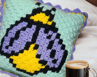 May include: A square crochet pillow featuring a light mint green background with a central pixelated insect design in black, yellow, and lavender. The pillow is finished with a decorative lavender scalloped crochet border along all four edges.