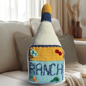 May include: A large, handmade crochet pillow shaped like a ranch dressing bottle. The pillow features a white body, a yellow and blue label with the word "RANCH" in blue, and decorative crochet details like a carrot and a flower.
