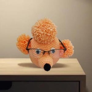 Może przedstawiać: Ręcznie robiona, brzoskwiniowa głowa pudla na szydełku z czarnym nosem, w okularach w czarnych oprawkach. Pudel ma puszysty pompon na czubku głowy i dwa mniejsze pompony po bokach. Pudel siedzi na drewnianej powierzchni.