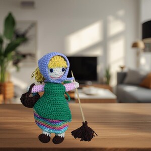 May include: A handmade crochet doll depicting a woman with blonde hair, wearing a blue headscarf, green dress, and a colorful skirt. The doll holds a broom and a brown bag. The doll is approximately 7 inches tall.