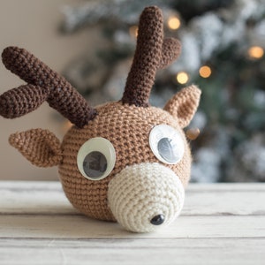 May include: A brown crocheted reindeer head with large white eyes and a white crocheted nose. The reindeer is sitting on a white wooden surface.