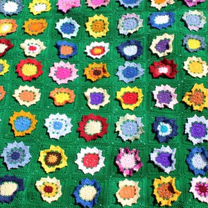 May include: A handmade, green crocheted blanket featuring numerous colorful flower motifs. The flowers are in various shades of pink, yellow, blue, and purple, creating a vibrant and textured pattern. The blanket is a cozy, handcrafted item.