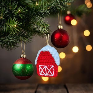 May include: Christmas tree ornaments, including a red and green bauble, a red crocheted barn with a white roof, and a solid red bauble. The ornaments are hanging from a Christmas tree branch with blurred lights in the background.