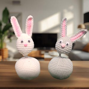 May include: Two handmade crochet bunny figures. One is white with pink ear lining and a pink nose, the other is gray with pink ear lining and a pink nose. Both have googly eyes and sit atop a white or gray ball.