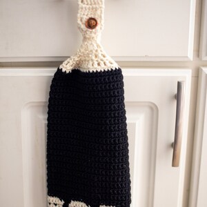May include: A black and white crocheted kitchen towel with a wooden button hanging on a white cabinet door.