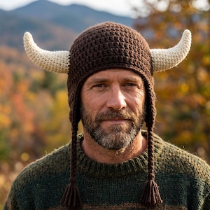 May include: A brown crocheted hat with cream-colored horns and ear flaps. The hat has a textured pattern and is worn by a person. The background is blurred, with autumn foliage and a mountain.
