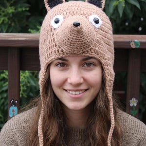 May include: A tan crochet hat featuring a fox design with pointed ears, a protruding snout, and two white eyes with black pupils. The hat includes long braided earflaps with tassels at the ends, crafted from light brown yarn with black accents.